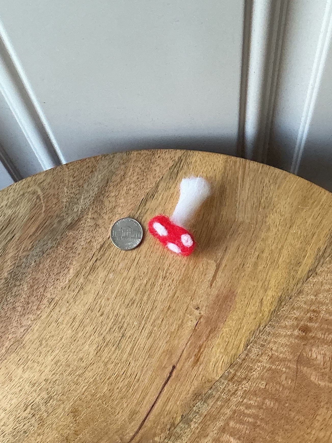 A round wooden side table viewed from above, with a purple and white patterned fabric draped over the left edge and a small red-and-white mushroom-shaped object and a coin on the tabletop. Part of a paneled white door is visible on the right.