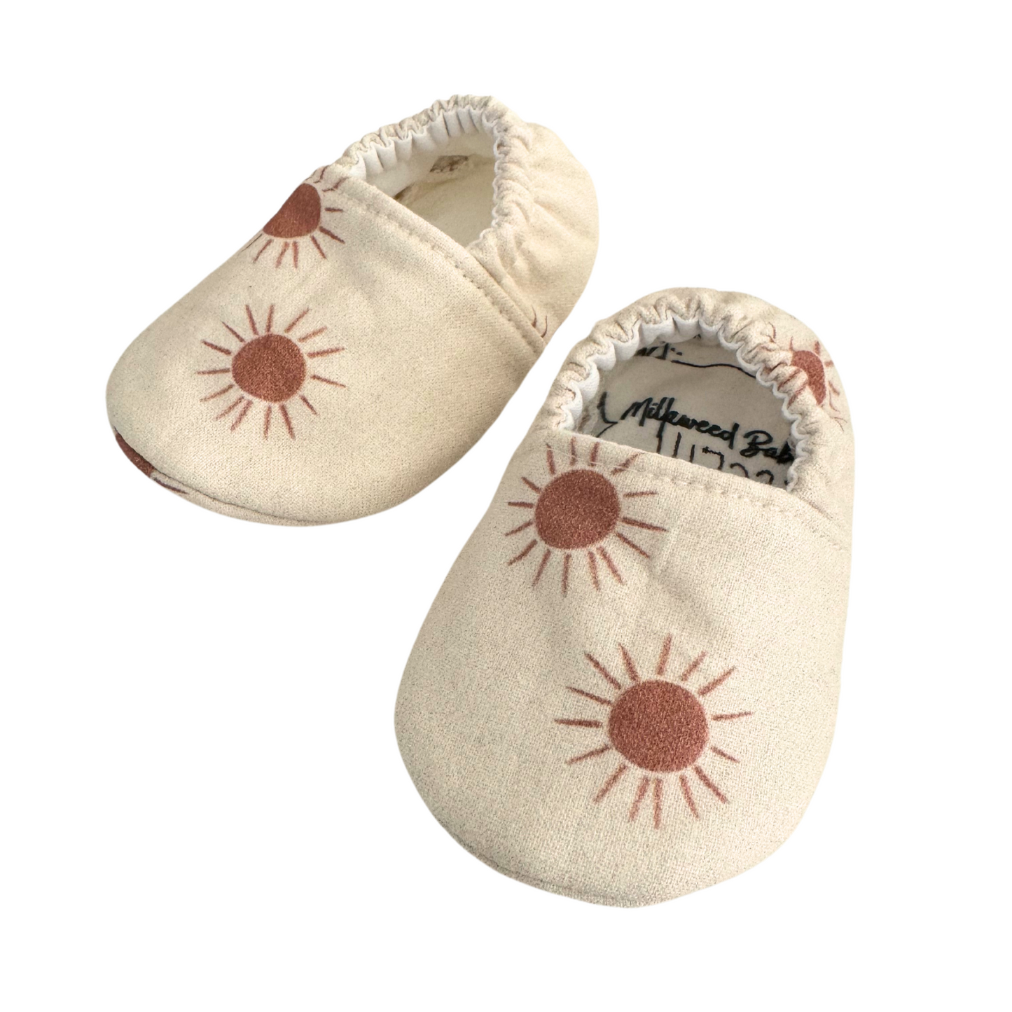 Baby moccasins with a neutral beige base featuring rust-colored sunburst prints, handmade with vegan materials and an elastic back for a snug fit, size 0-3 months, ready to ship.