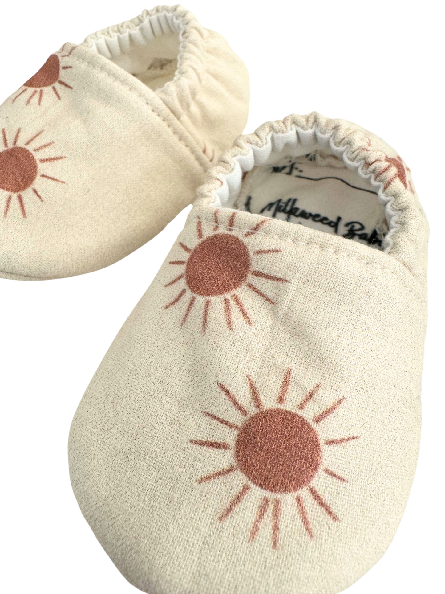 Baby moccasins with a neutral beige base featuring rust-colored sunburst prints, handmade with vegan materials and an elastic back for a snug fit, size 0-3 months, ready to ship.