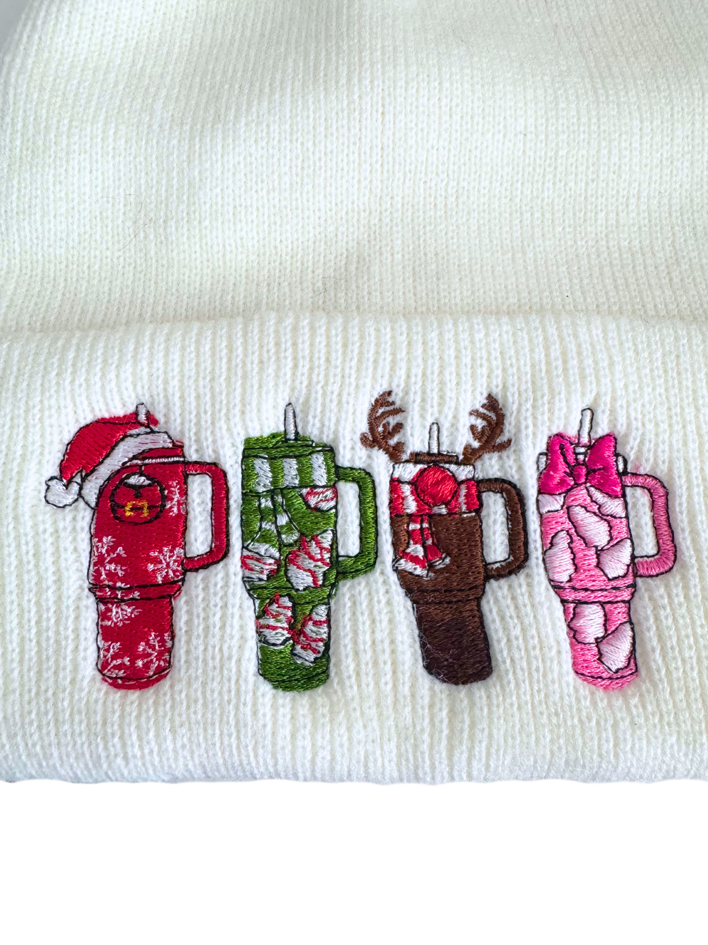 White beanie with embroidered holiday-themed Stanley-style cups, including designs of Santa, reindeer, candy canes, and marshmallows.