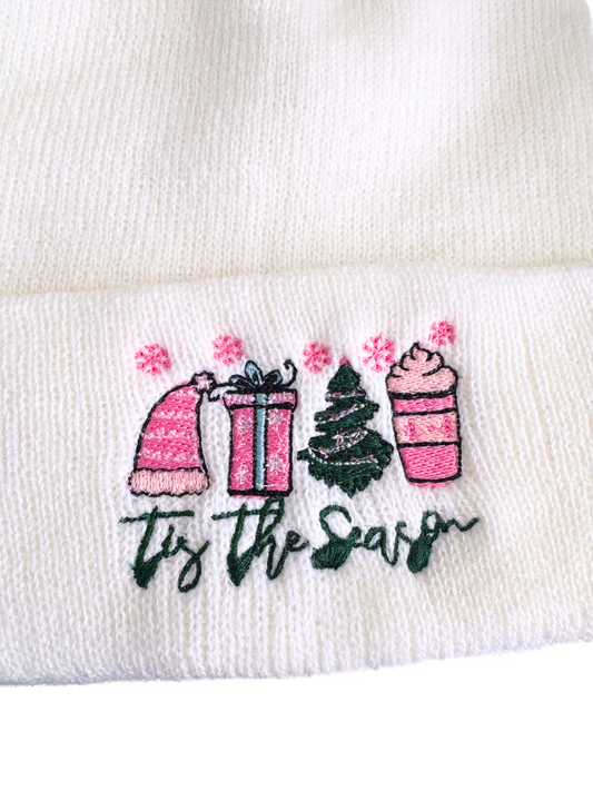 White beanie with "Tis The Season" festive embroidery featuring a pink hat, gift box, Christmas tree, and hot cocoa cup.