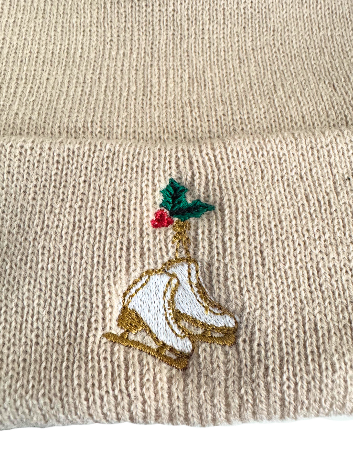 Beige knit beanie with embroidered golden ice skates and holly design.