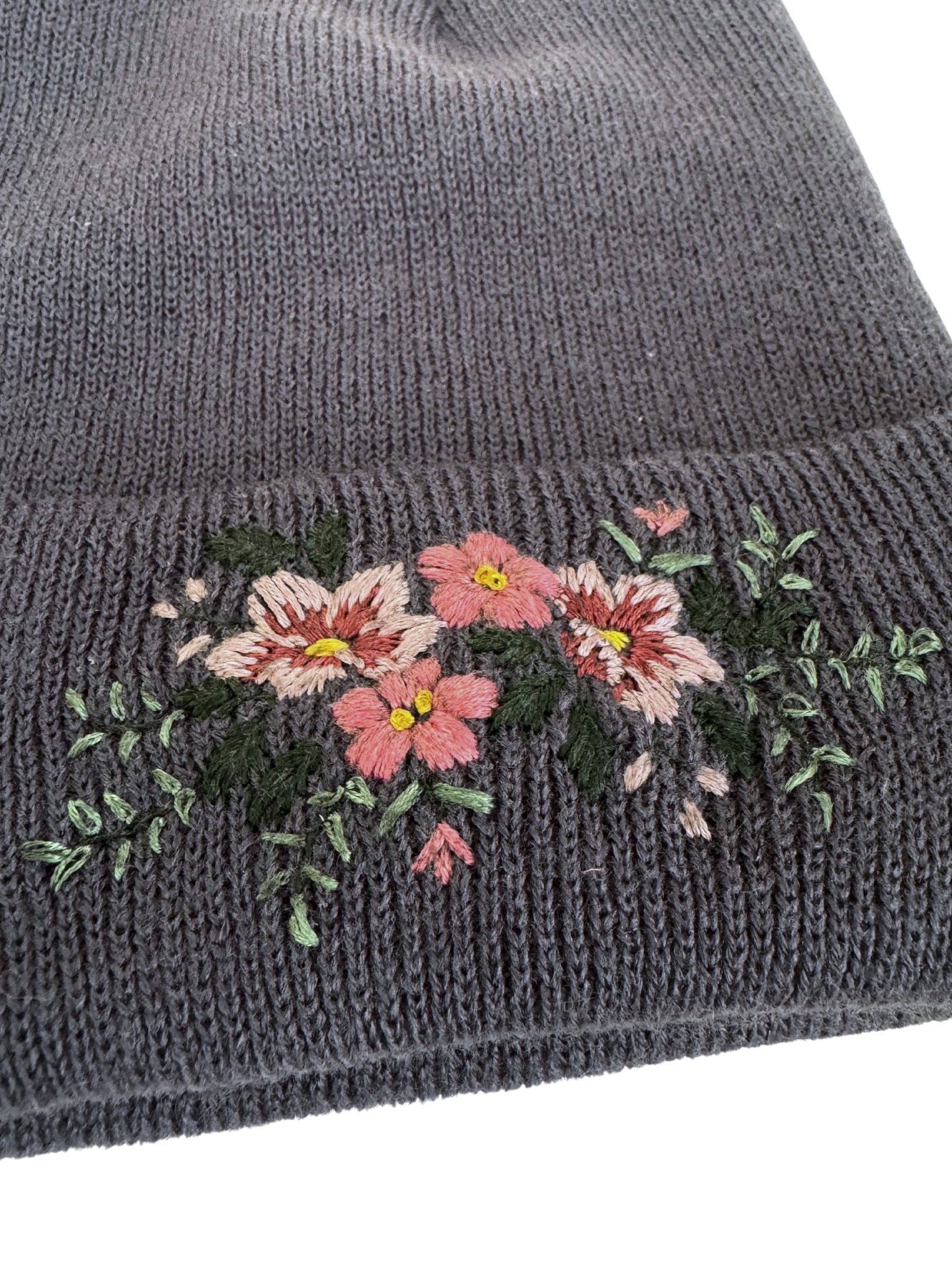 Close-up of hand-embroidered floral design on a blue/gray knit beanie, showcasing intricate craftsmanship.