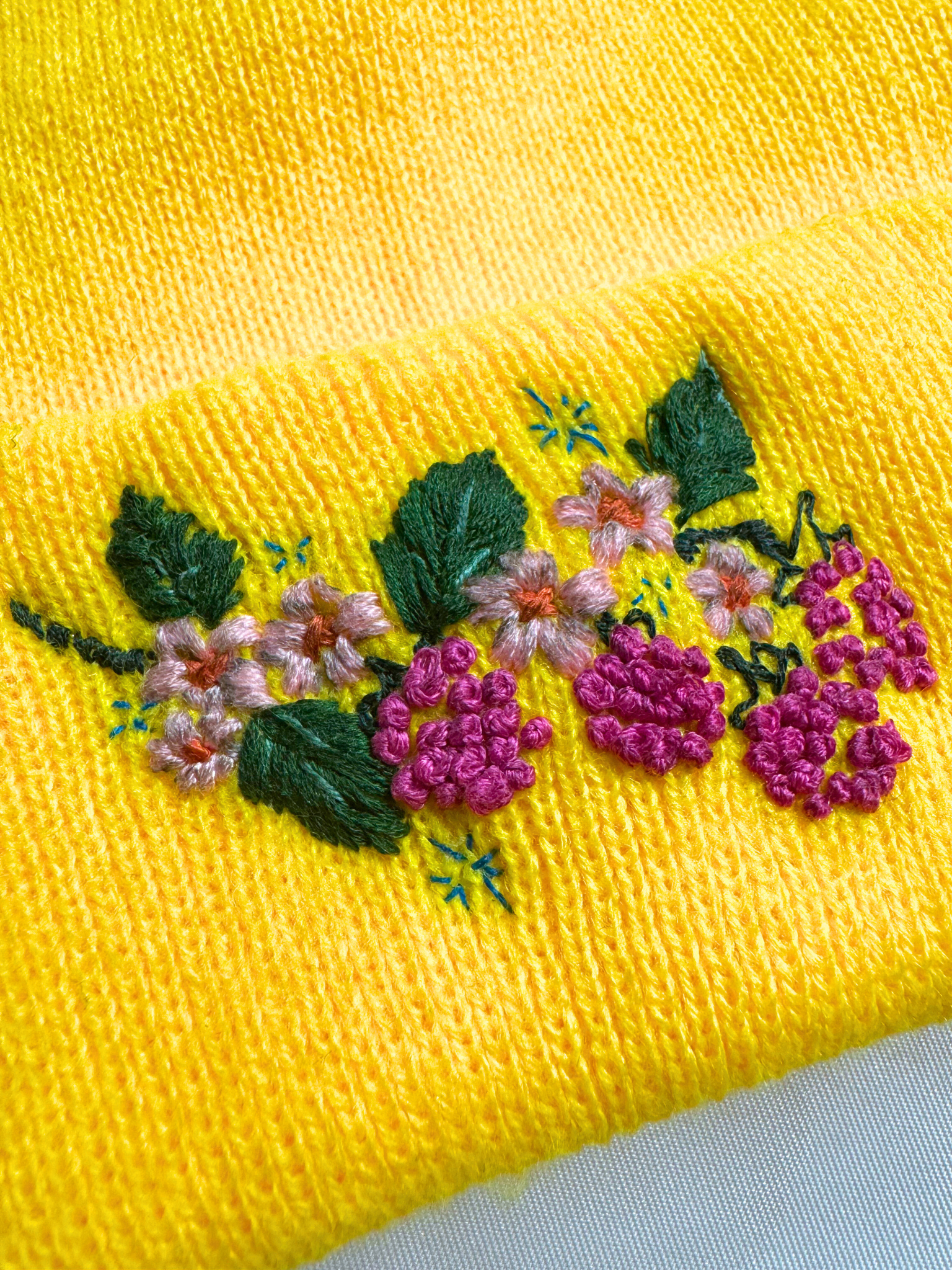 Close-up of embroidered berry and floral design on a yellow knit beanie, highlighting vibrant colors and intricate stitching.