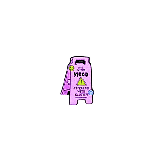 Close-up of enamel pin shaped like a pink caution sign reading “Not in the Mood / Approach With Caution”
