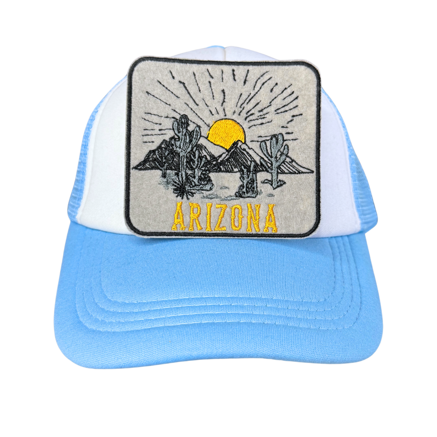 "Blue and white trucker hat with an embroidered Arizona desert sunset patch featuring cacti, mountains, and a setting sun."