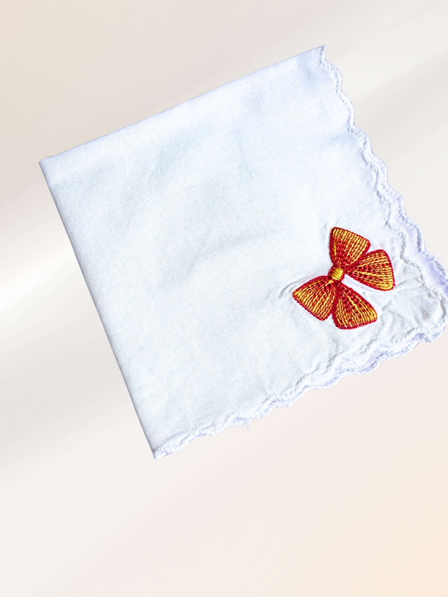 A folded embroidered handkerchief showcasing a striking red and gold bow stitched onto soft white fabric, handmade in Kansas City.