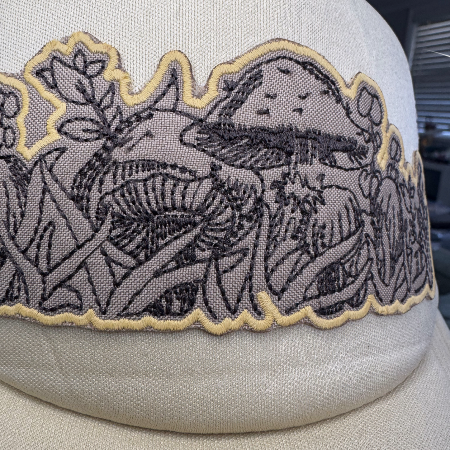 Closeup of scalloped black and gold mushroom embroidery on neutral foam-front trucker hat