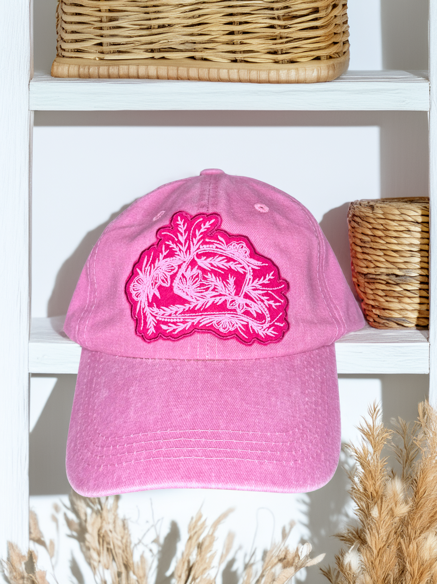 Pink cap with white floral design on a white shelf with wicker baskets and plants.