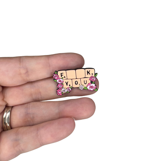 Enamel pin spelling out 'F*ck You' shown in hand for scale, Scrabble-style with roses