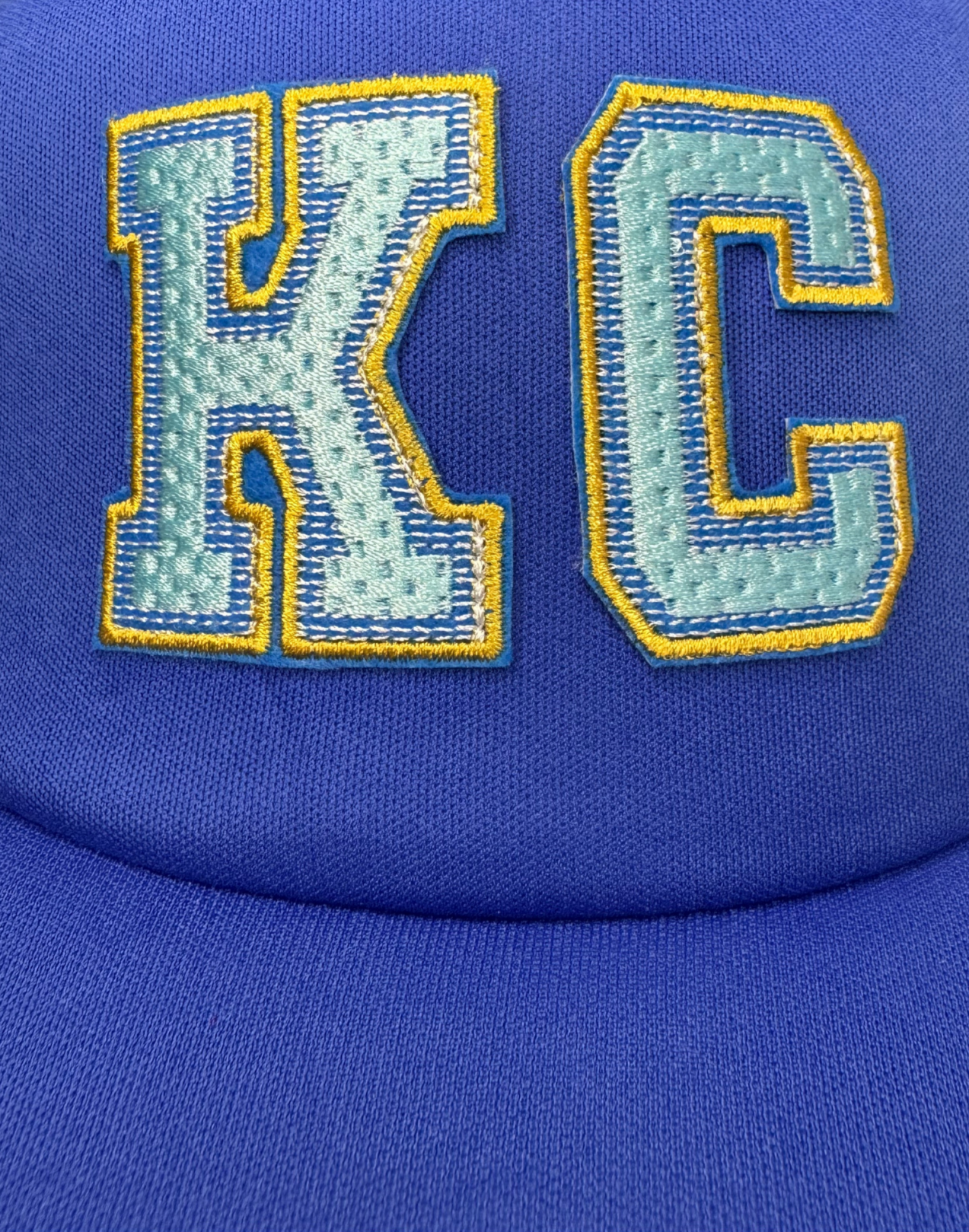 Closeup of varsity-style KC patch in light blue with gold outline on royal blue hat