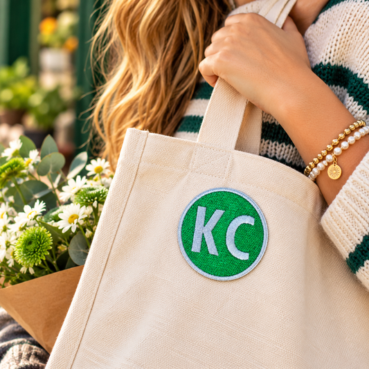 Green and white circular KC embroidered patch attached to a cream canvas tote carried by a person in a green striped sweater