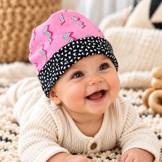 Smiling baby wearing pink lightning bolt beanie with black-and-white triangle cuff during supervised tummy time