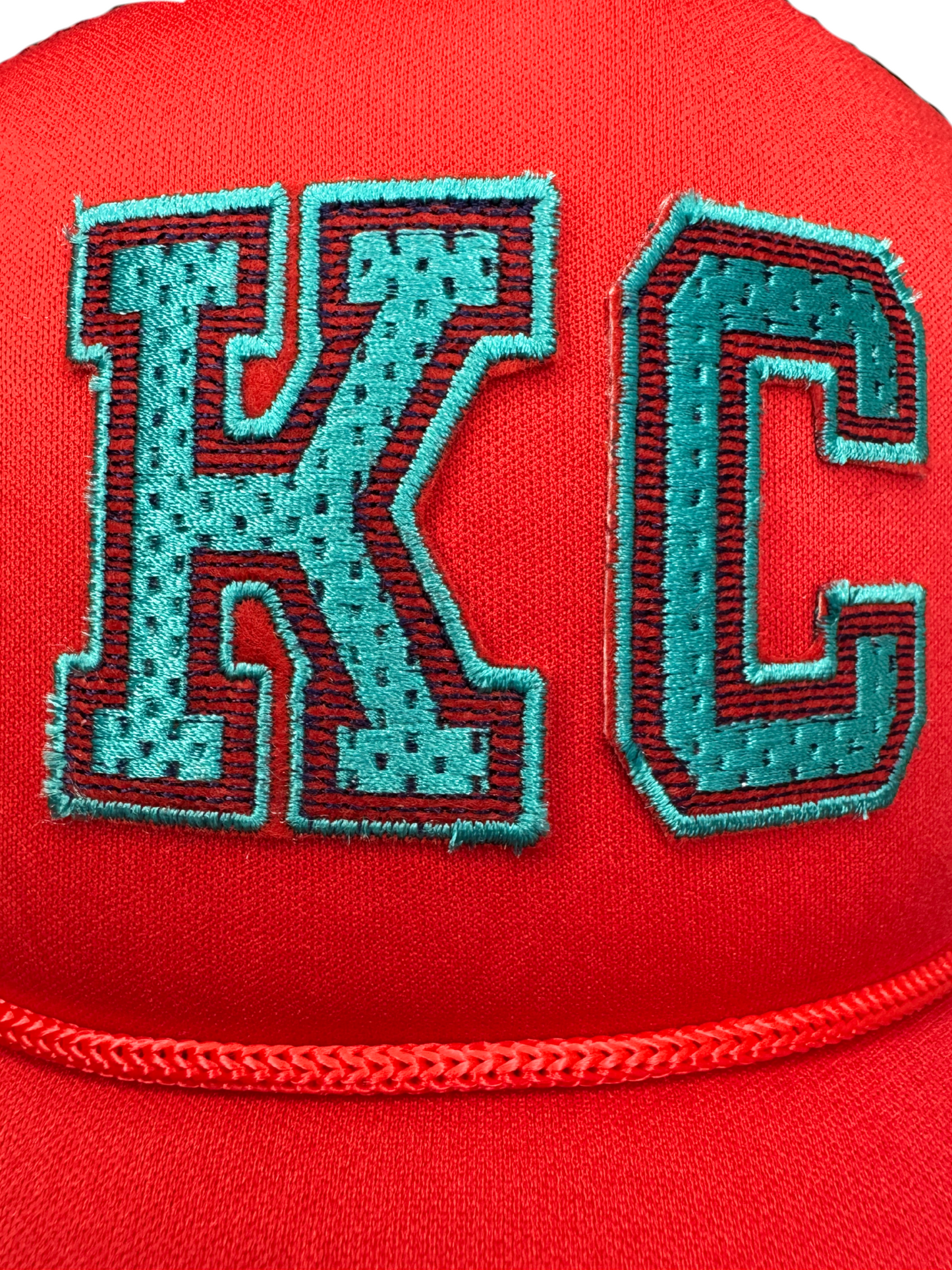 Closeup of varsity-style KC patch in teal embroidery on red trucker hat