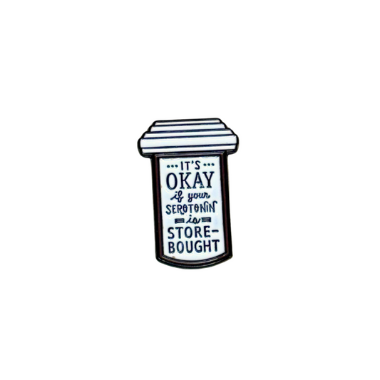 Black and white enamel pin shaped like a prescription bottle with text that reads “It’s okay if your serotonin is store-bought”
