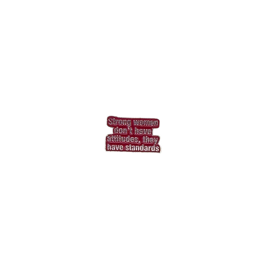 Hand holding a red and white enamel pin that says "Strong women don’t have attitudes, they have standards"