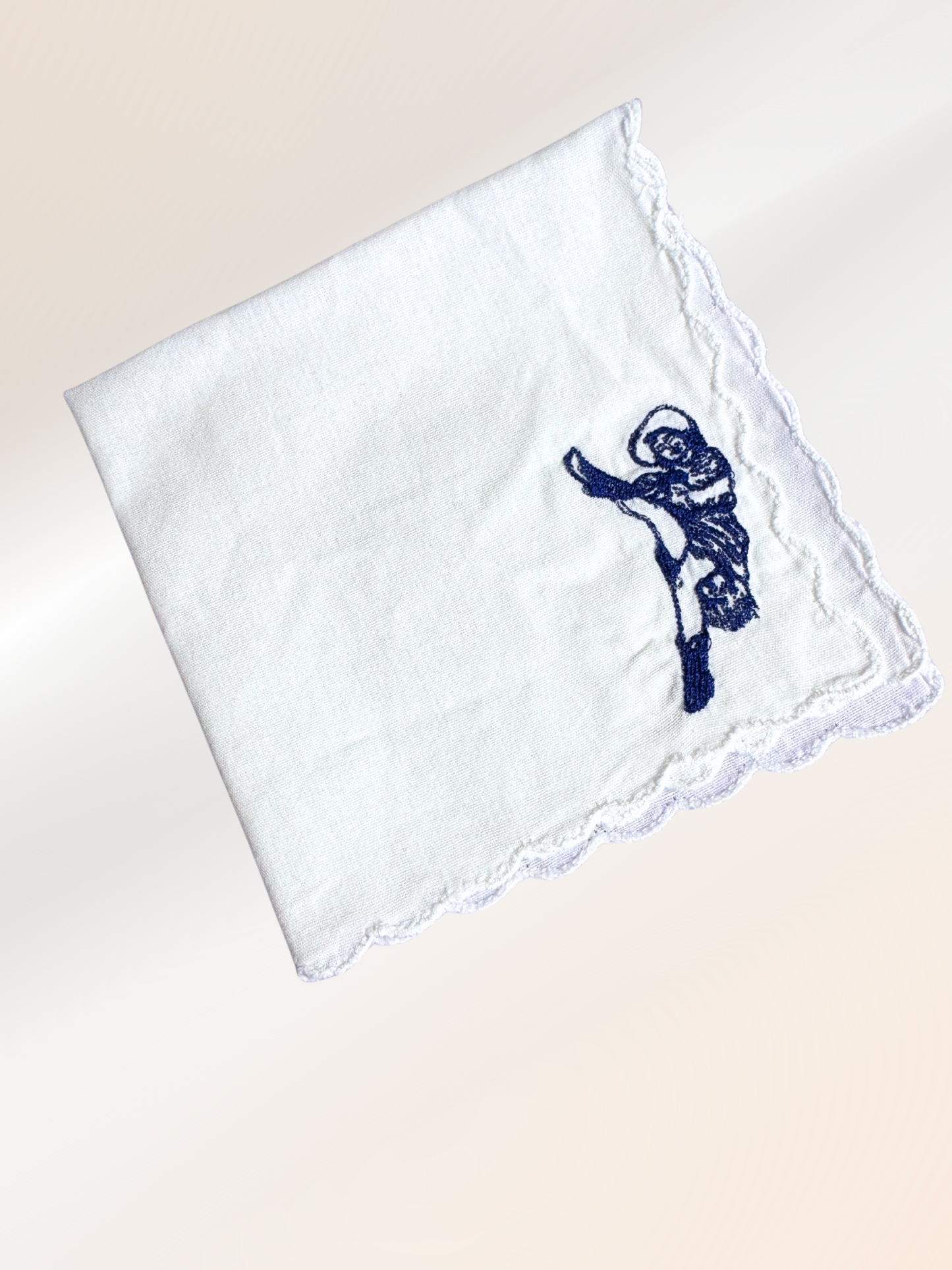 A folded embroidered handkerchief showcasing a vintage French cabaret dancer stitched in blue on soft white fabric, handmade in Kansas City.