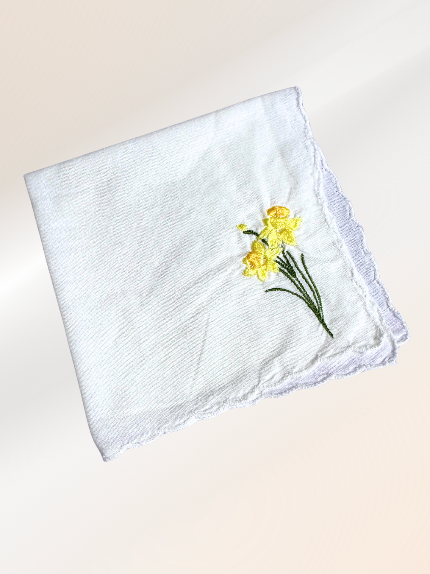 A folded embroidered handkerchief showcasing a delicate yellow daffodil design stitched onto soft white fabric, handmade in Kansas City.