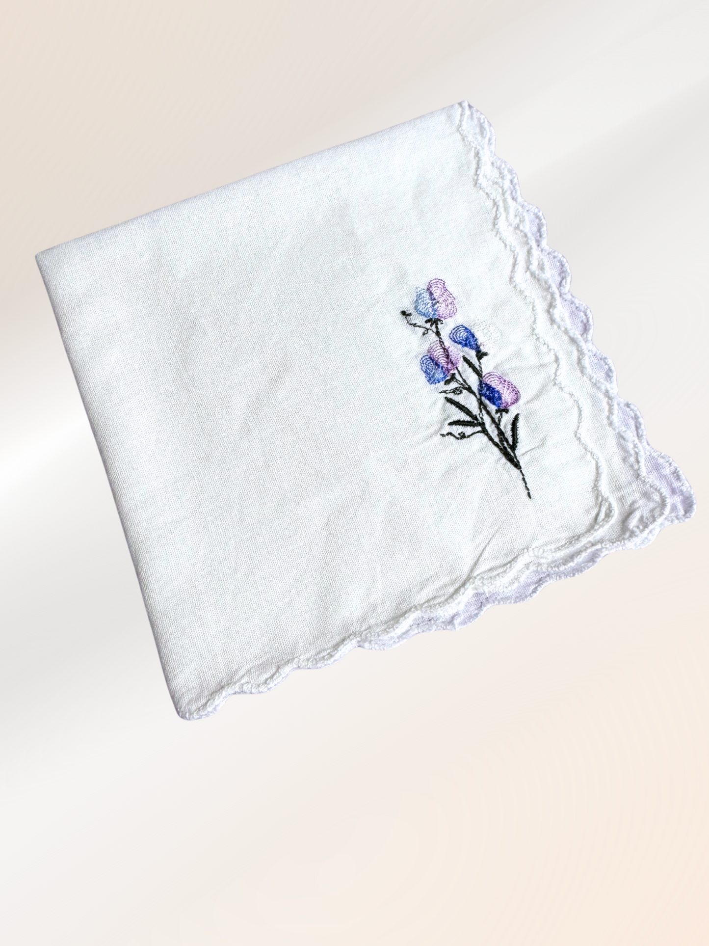 A folded embroidered handkerchief showcasing a delicate sweet pea vine stitched in lavender, blue, and green on soft white fabric, handmade in Kansas City.