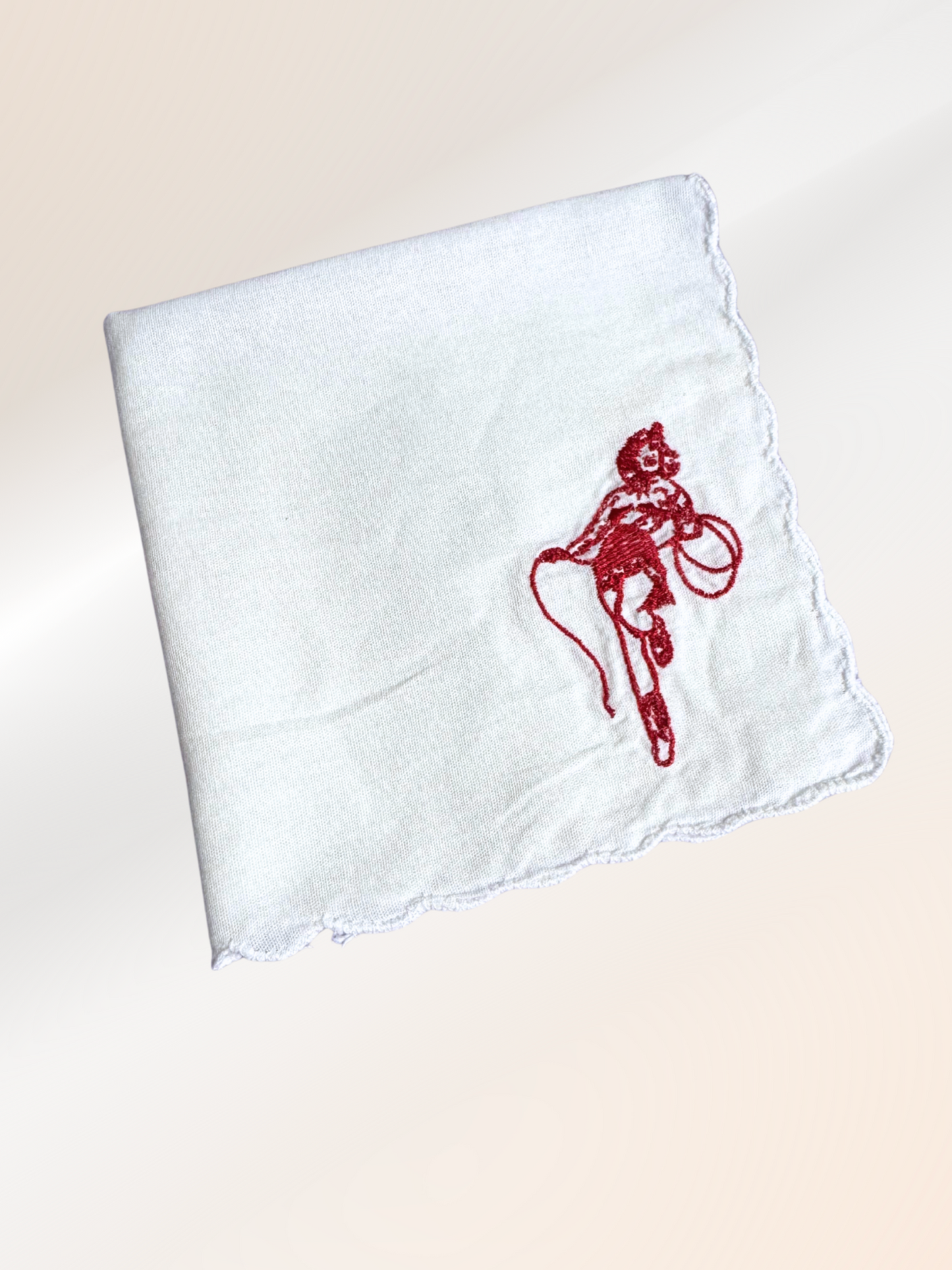 A folded embroidered handkerchief showcasing a retro cowgirl roper design in red on soft white fabric, handmade in Kansas City.