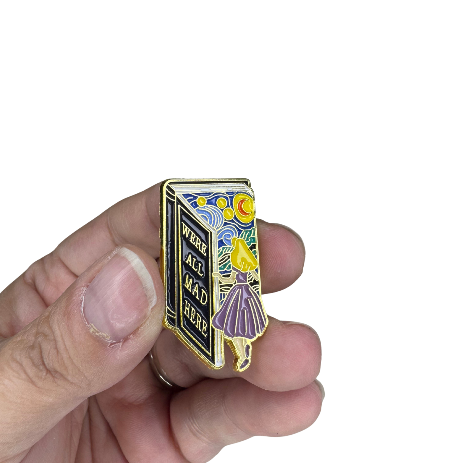 “We’re All Mad Here” enamel pin shown held in hand for size reference