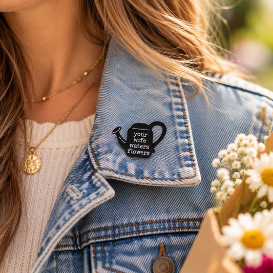 Taylor Swift-inspired your wife waters flowers black watering can enamel pin worn on a blue denim jacket lapel.