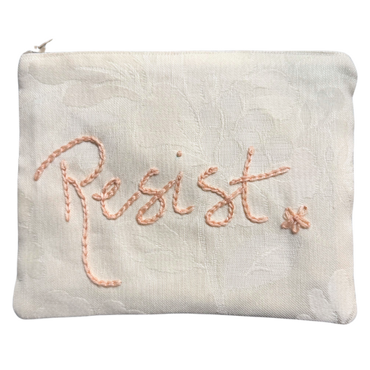 Hand-embroidered cream clutch with the word “Resist” in peach thread
