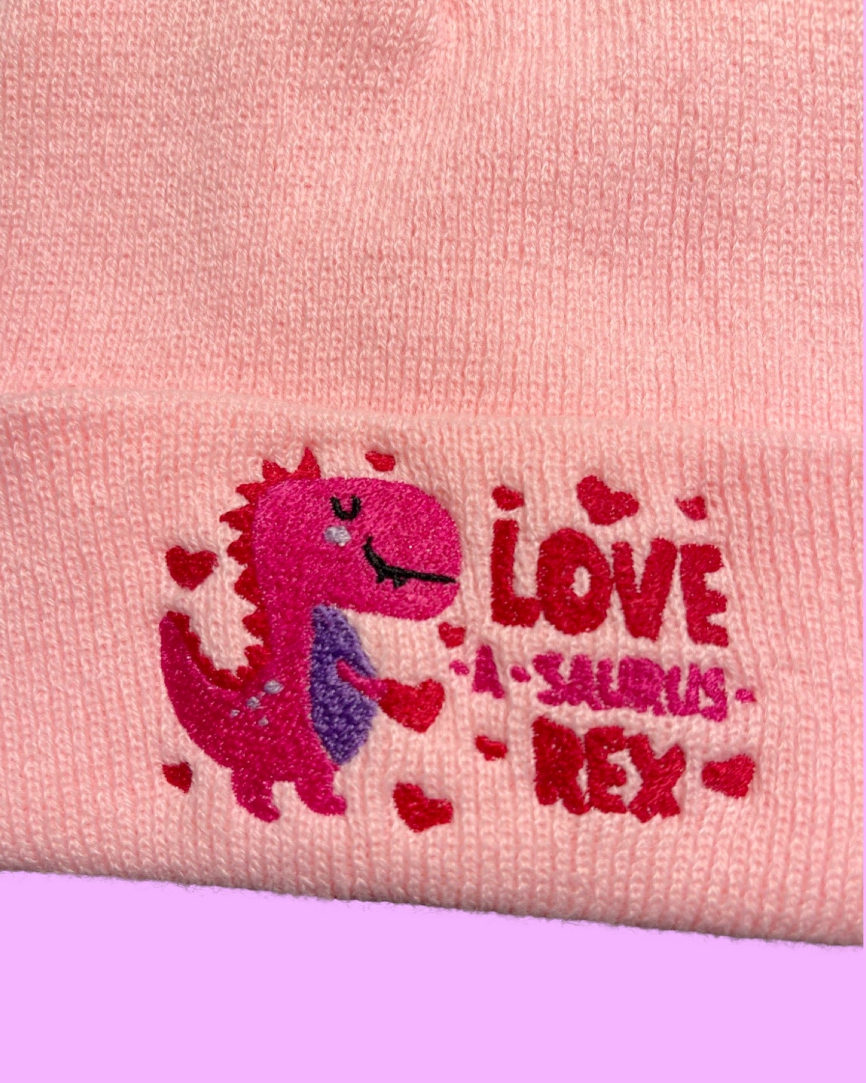 detail view light pink knit beanie embroidered with a pink red and purple dinosaur surrounded by hearts and the text Love-a-saurus Rex