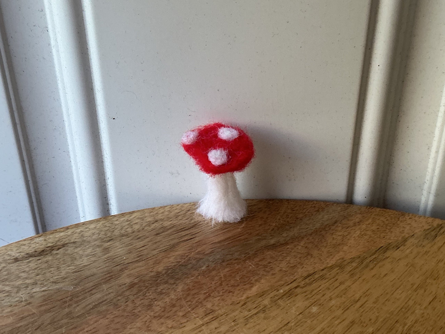 A small red and white felt mushroom decoration hanging from the edge of a round wooden board, shown against a white paneled background.