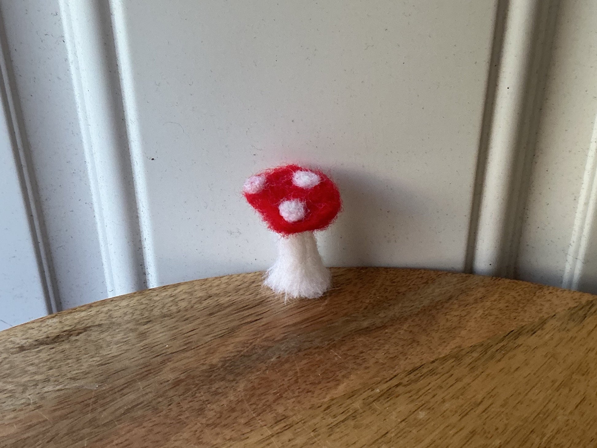 A small red and white felt mushroom decoration hanging from the edge of a round wooden board, shown against a white paneled background.