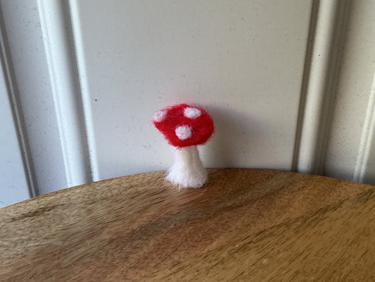 A small red and white felt mushroom decoration hanging from the edge of a round wooden board, shown against a white paneled background.