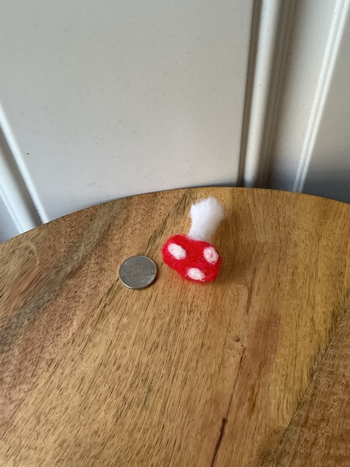 A small red and white mushroom-shaped object with a white fluffy top sits on a round wooden table next to a coin, with a white paneled wall in the background.