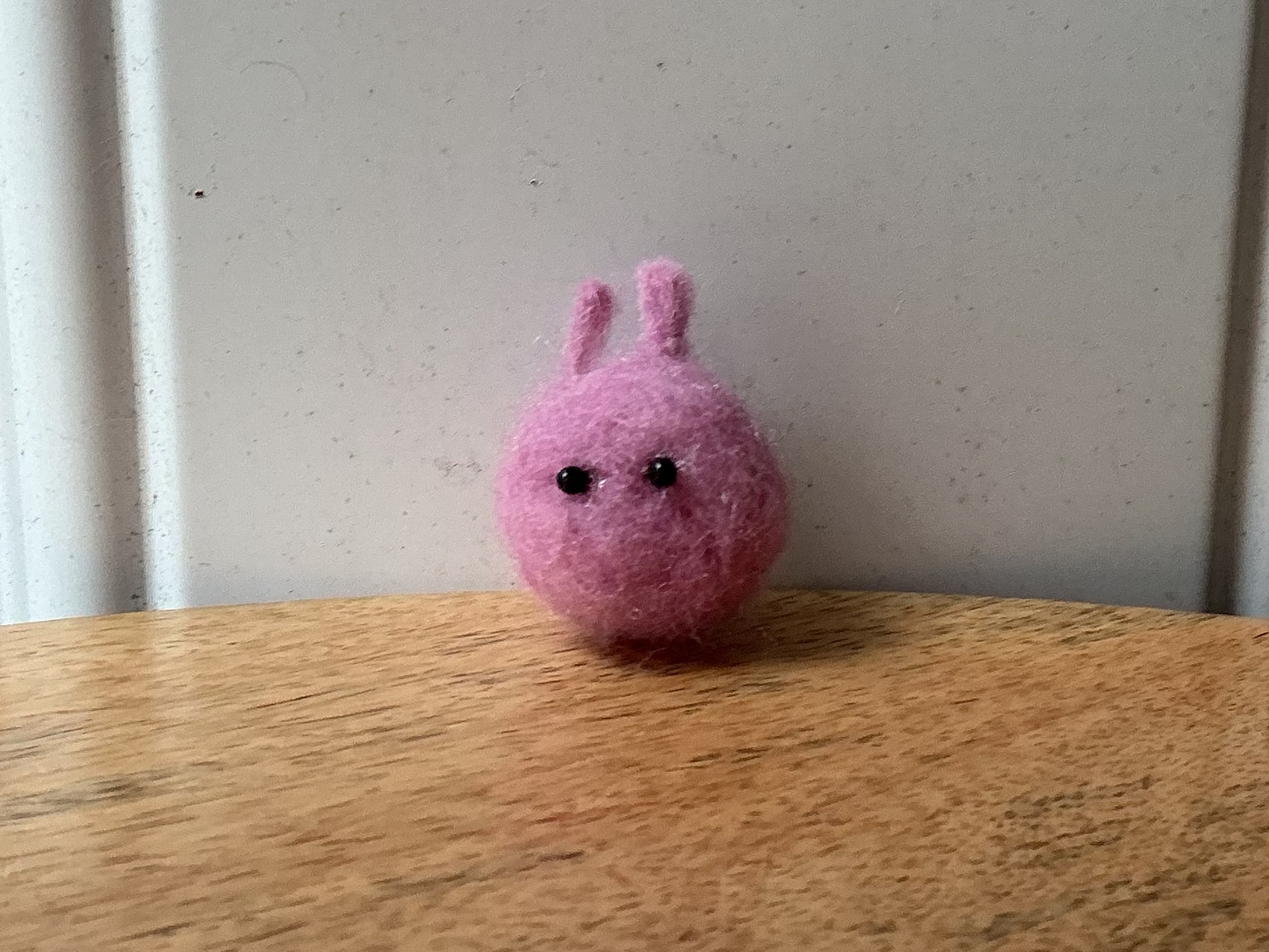 A small pink felt bunny with black embroidered eyes sits on a wooden tabletop against a white paneled wall.