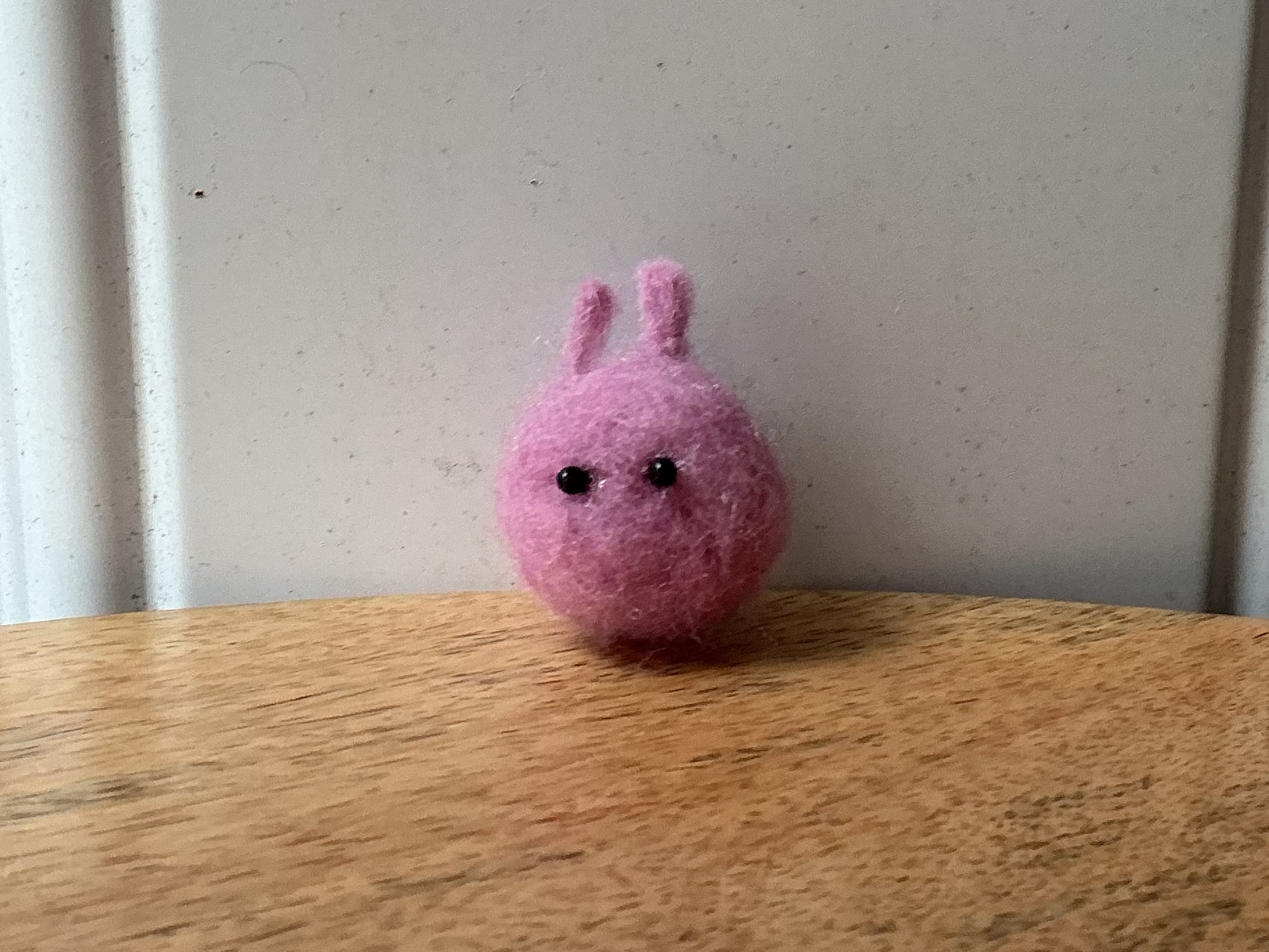 A small pink felt bunny with black embroidered eyes sits on a wooden tabletop against a white paneled wall.