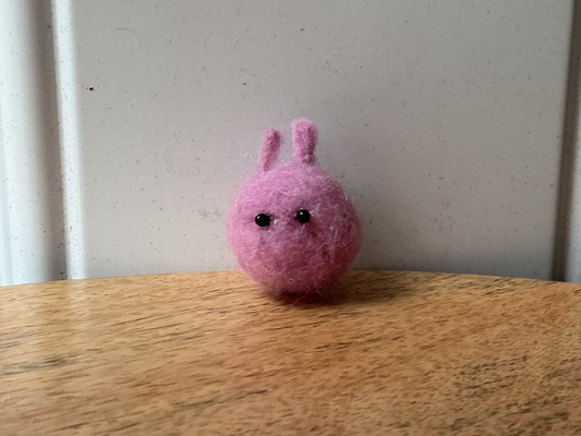 A small pink felt bunny with black embroidered eyes sits on a wooden tabletop against a white paneled wall.