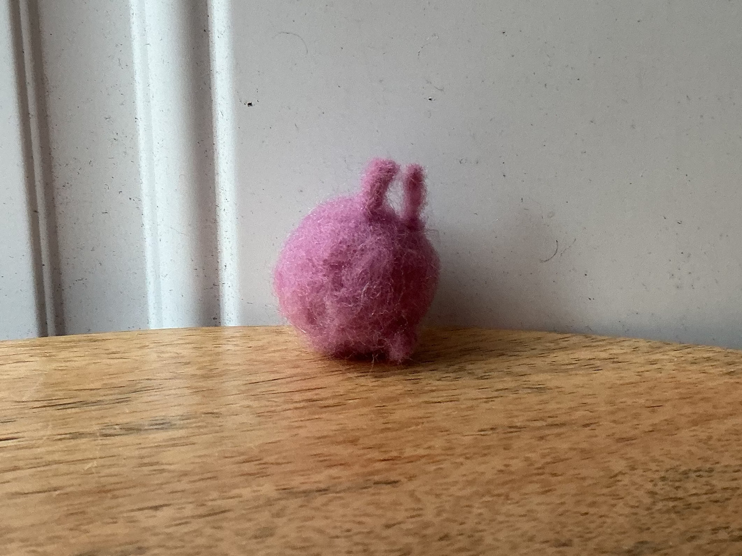 A small pink felted wool bunny figurine sitting on a wooden tabletop against a white paneled wall.