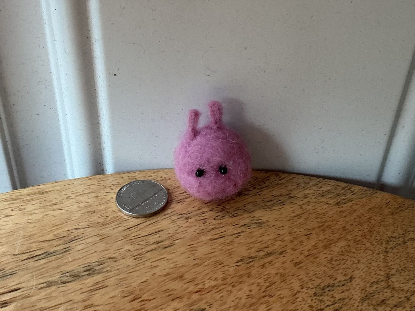 A small pink felt creature with two antennae and a simple face sits on a wooden tabletop next to a coin for scale, with a white paneled wall in the background.