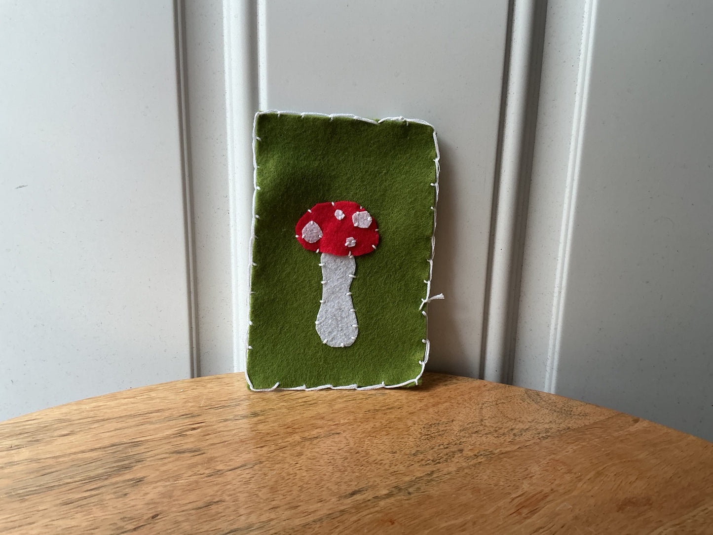 A handmade felt wall hanging with a green background and a red and white mushroom applique, standing on a wooden table against a white paneled wall.