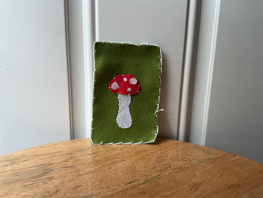 A handmade felt wall hanging with a green background and a red and white mushroom applique, standing on a wooden table against a white paneled wall.