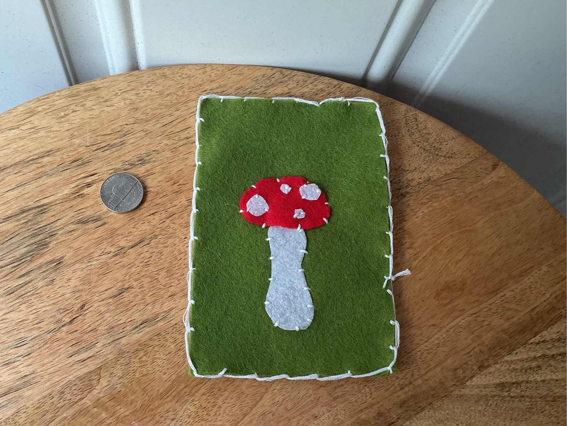 A handmade felt wall hanging featuring a red and white mushroom design on a green background, placed on a wooden table next to a coin for scale.