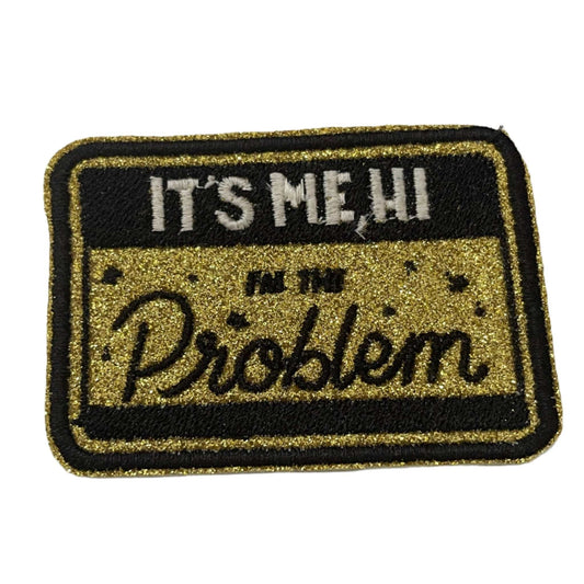 Handmade iron-on patch with Taylor Swift lyrics "It's Me, Hi, I'm the Problem" on glittery gold background with black outline.