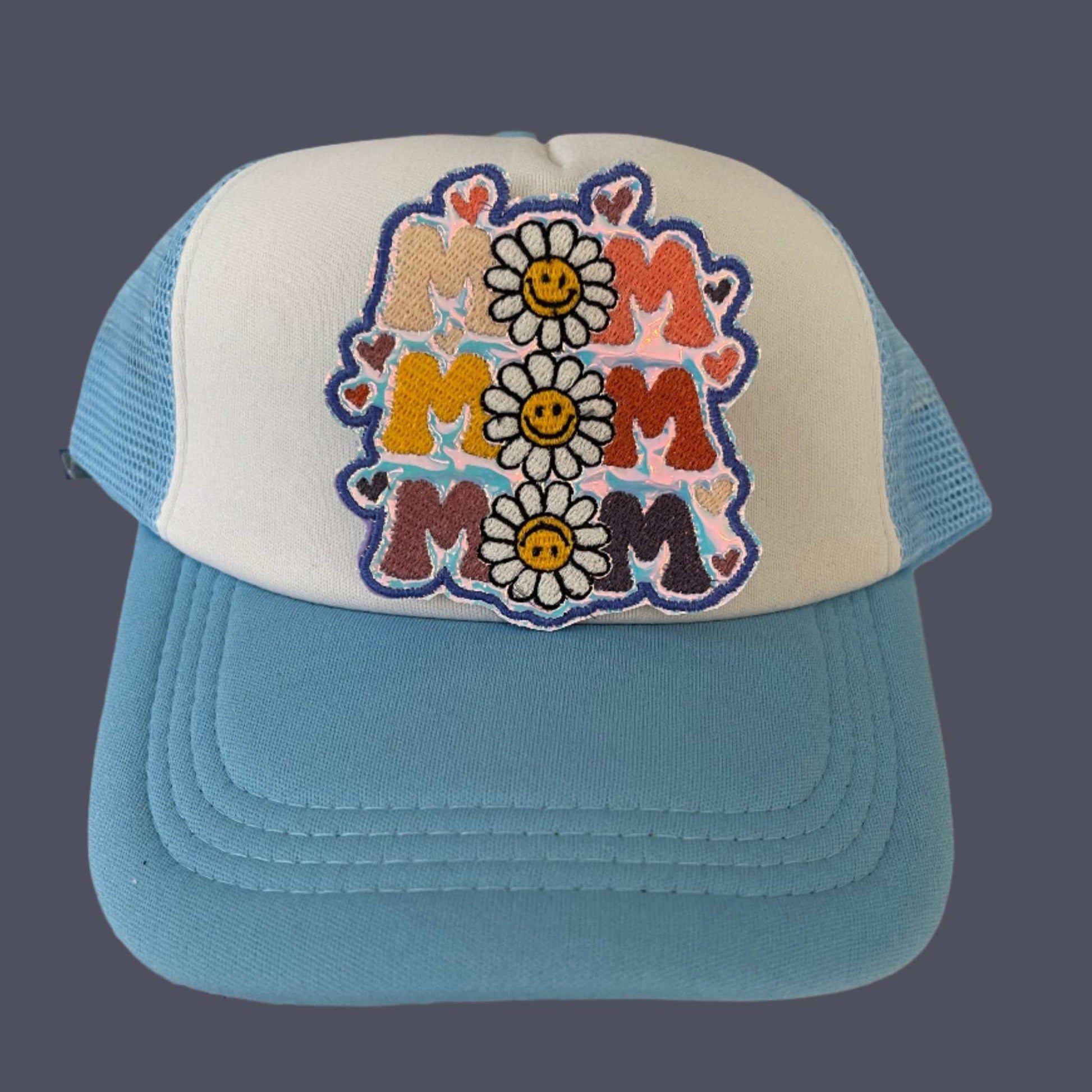 Iron-on patch featuring the word "MAMA" repeated in beige, pink, orange, red, and purple with small hearts and two smiling daisies, outlined in dark blue thread.