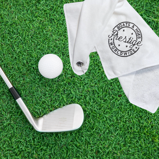 Prestige Worldwide 'Boats n Hoes' Microfiber Golf Towel on grass with golf club and ball.