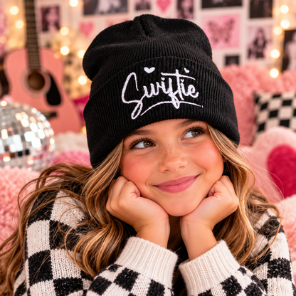 Young girl wearing the black Swiftie embroidered youth beanie with white script and heart details.