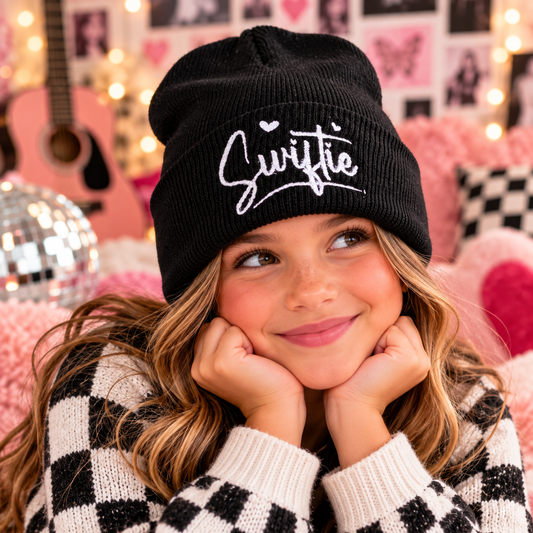 Young girl wearing the black Swiftie embroidered youth beanie with white script and heart details.