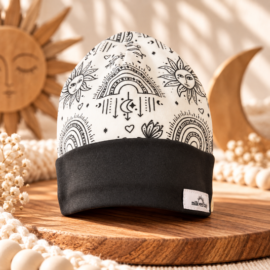 Cream organic cotton baby beanie with black sun moon rainbow print and charcoal cuff, styled on wood