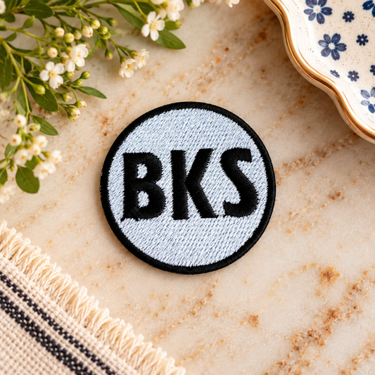 Black and white embroidered Brookside BKS round iron-on patch styled on warm stone with white flowers and vintage ceramic dish