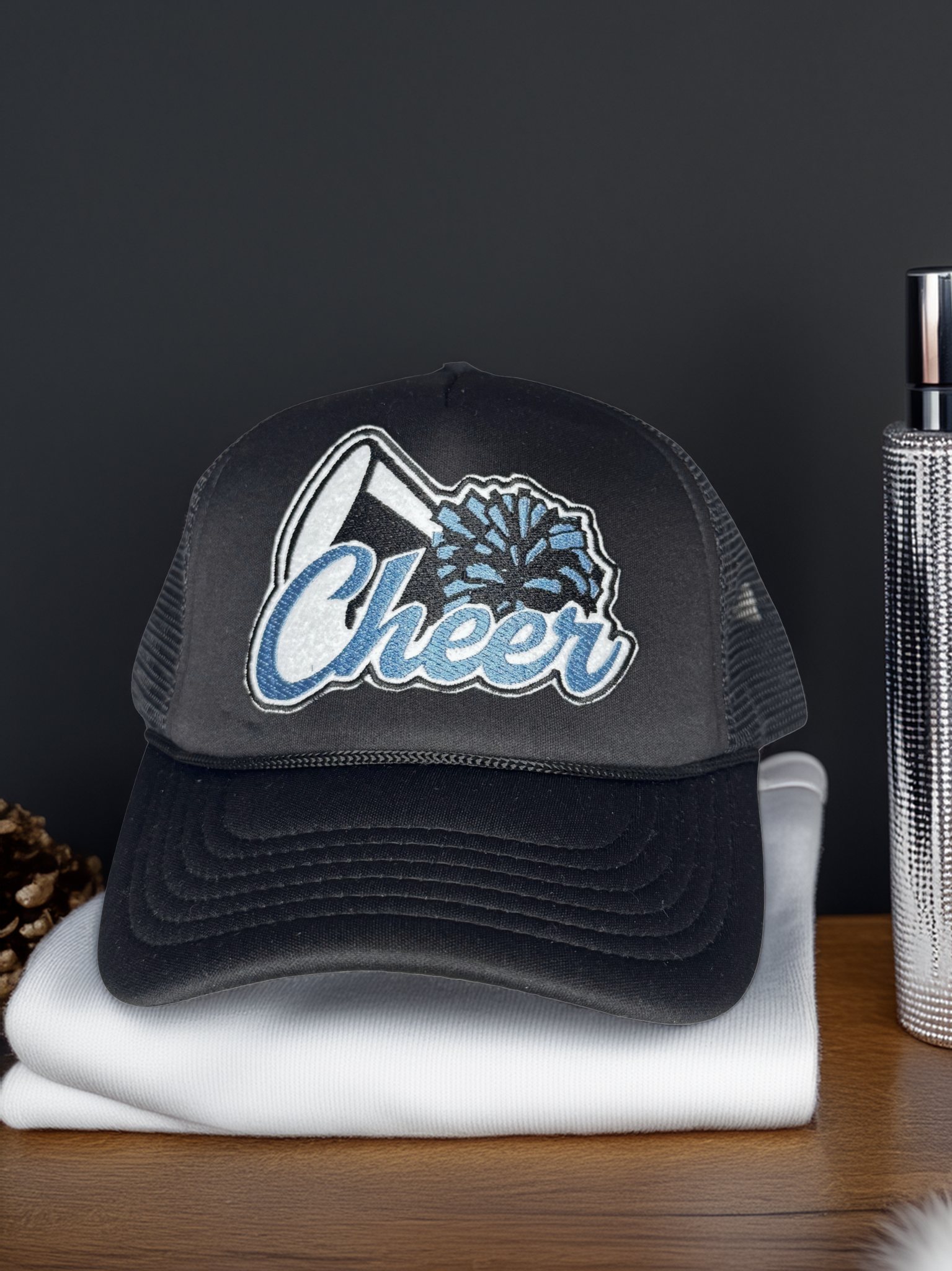 Black trucker hat with 'Cheer' and decorative elements on a white surface with a dark background