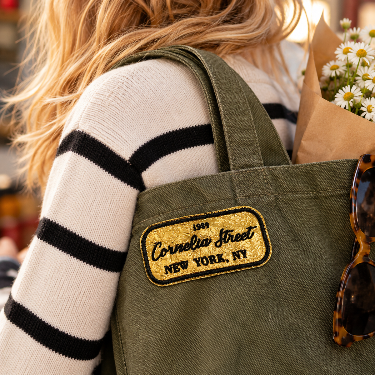 Cornelia Street gold and black embroidered patch on olive canvas tote carried over a striped sweater