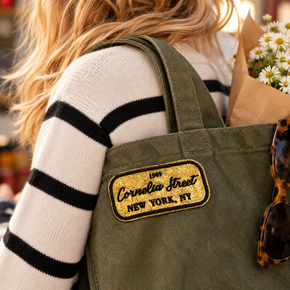 Cornelia Street gold and black embroidered patch on olive canvas tote carried over a striped sweater